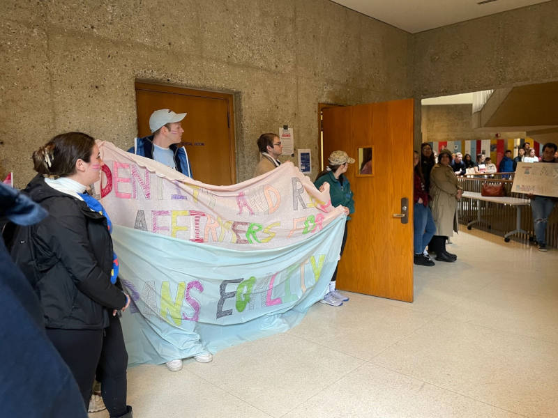 Protestors holding up "trans equality" fabric sign in lobby of Stanford Law School lecture hall in protest of Judge Kyle Duncan